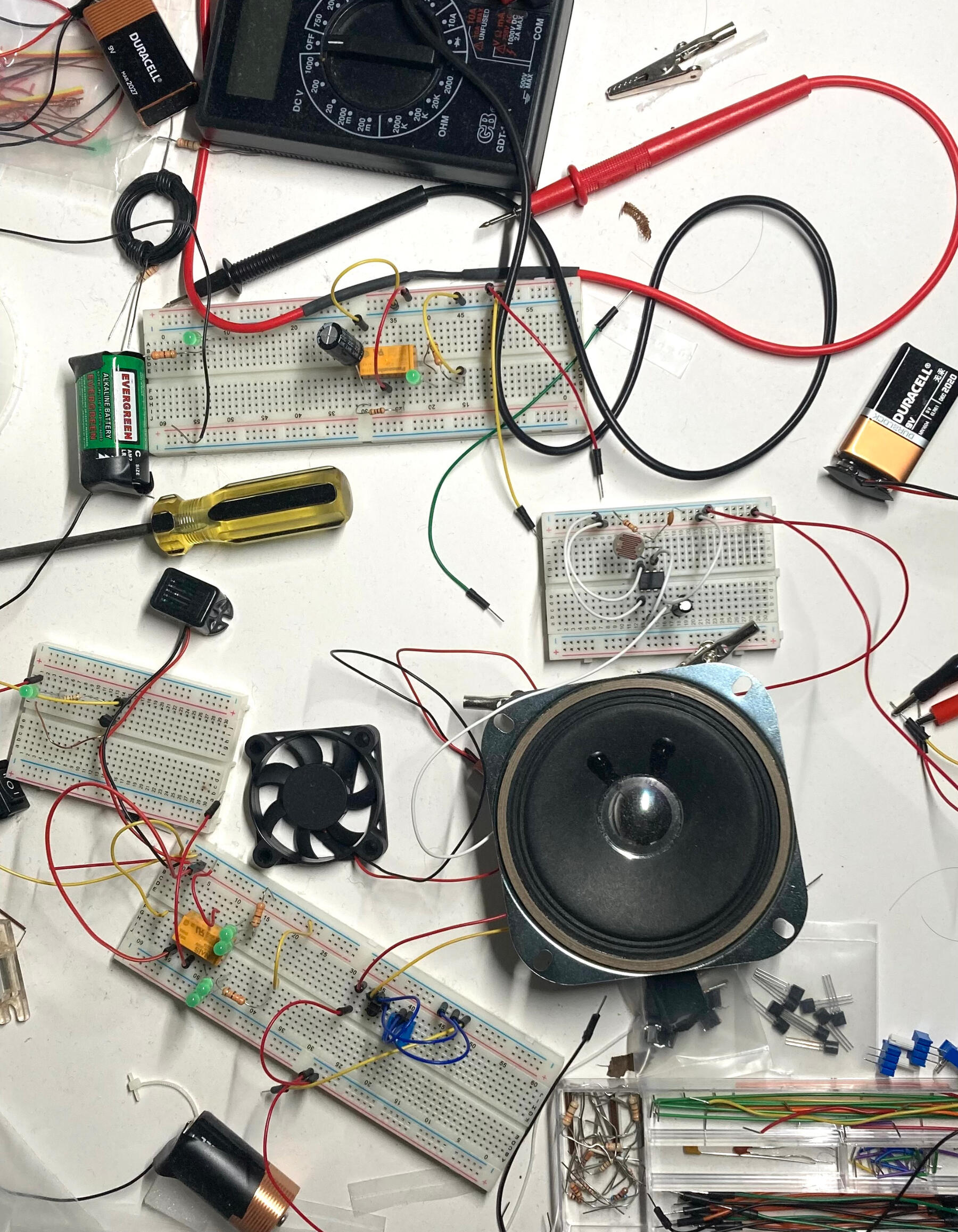Color photograph: Overhead view of white table top scattered with an array of electronics supplies: bread boards, jumper wires, LEDs, a speaker, a small fan, batteries, screwdriver, a multimeter etc.