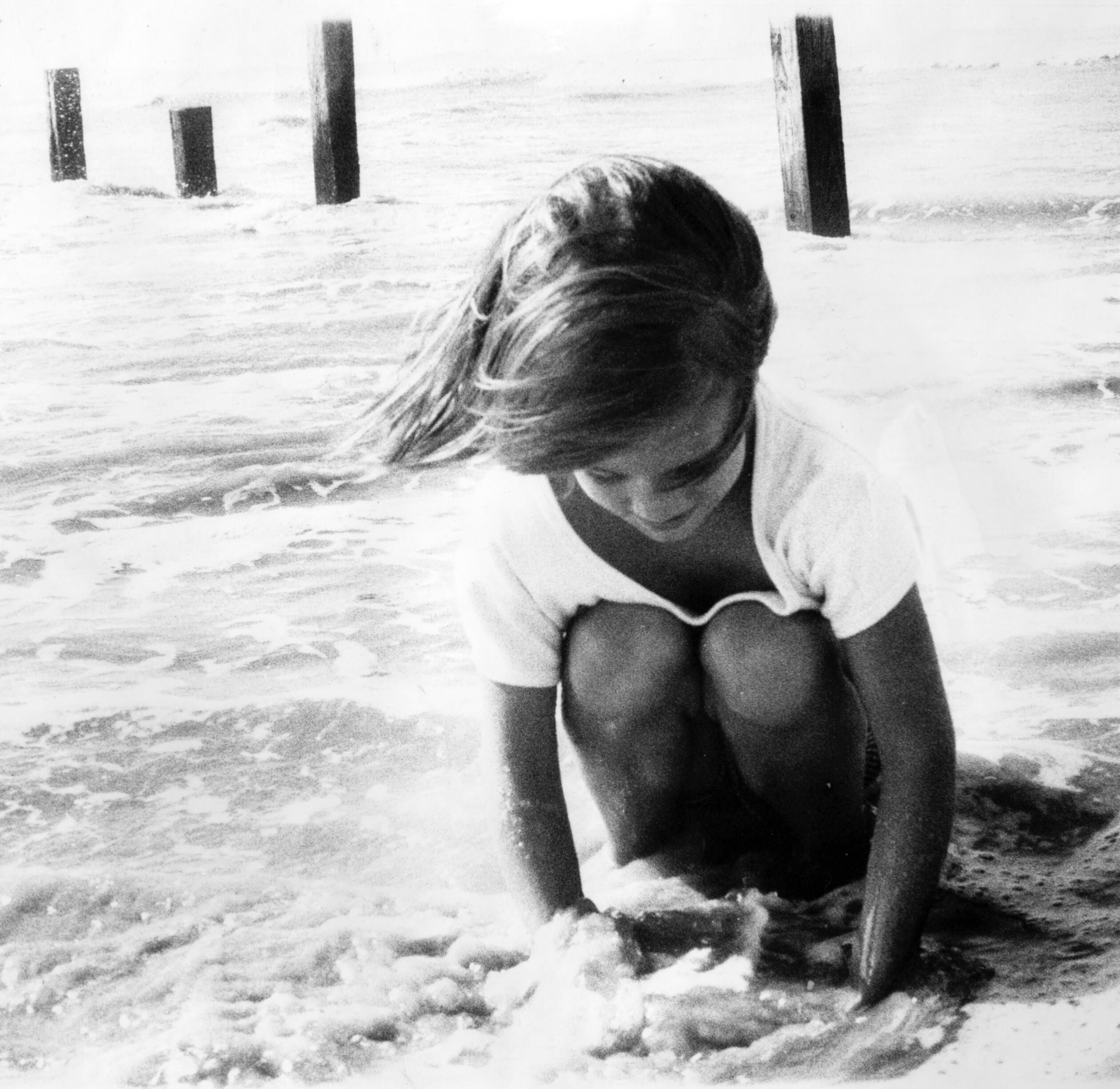 Black-and-white photograph: A white girl about five years old crouches in low surf, fascinated by the flow of water through her hands and feet.