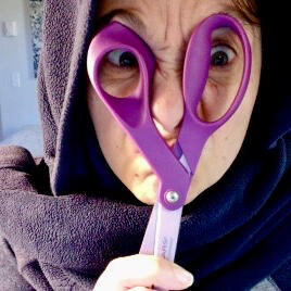 Color photograph: Close-up headshot with scrunched face looking through the handles of purple scissors. Blue fuzzy cloth draped around head and neck.