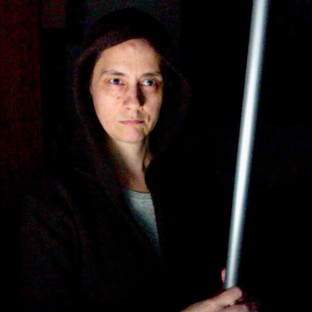 Color photograph: Imitation of Obi-Wan Kenobi with light saber. Serious face in stark light with cloak around head. Shining silver stick held in front.